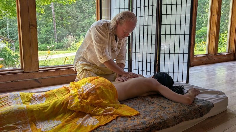 Sergio working on the upper back of a client lying face down by a window with a garden view, a yellow sarong over her legs